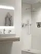 Dusche mit Kopfbrause und Handbrause von Ideal Standard. Helle Fliesen und ein modernes Waschbecken mit Holzregal sind im Raum vorhanden. Handtuch hängt neben dem Spiegel.
